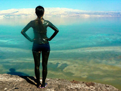 Woman covered in mud standing on shore of Dead Sea, with back to camera