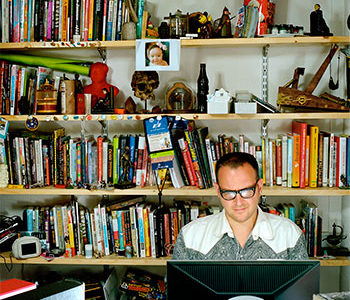 man on computer with wall of books behind him on shelves